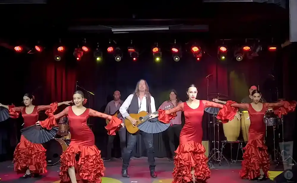 Flamenco dancers in red dresses perform with a guitarist and percussionists on a dimly lit stage.