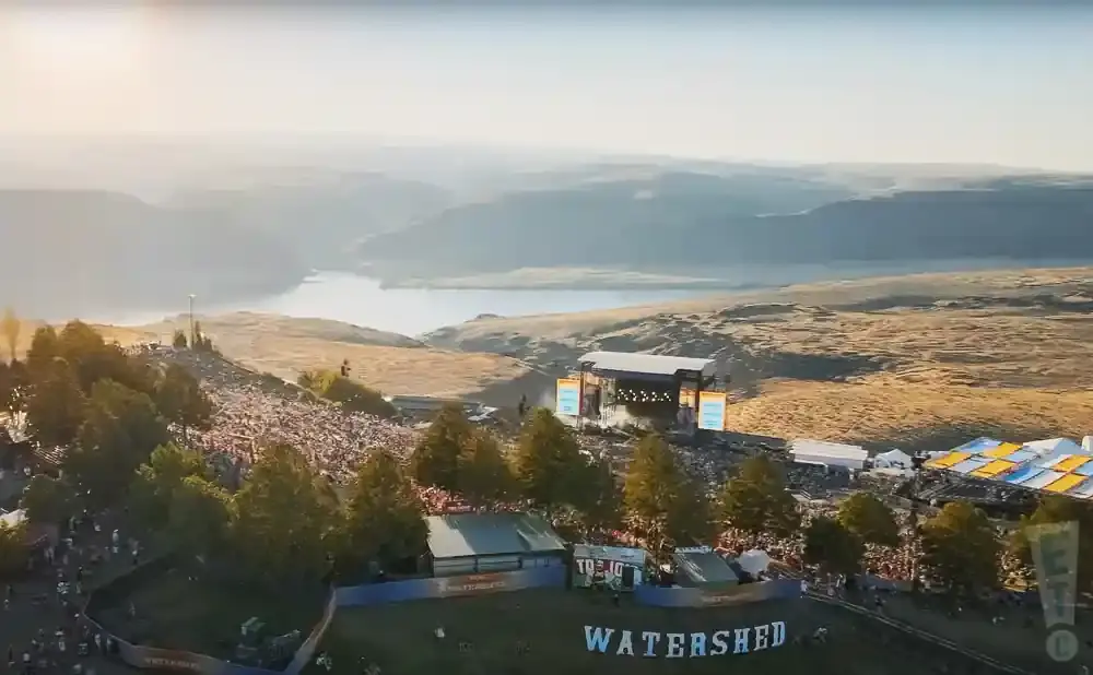 Aerial view of a large outdoor concert with a stage, a crowd, and tents, set against a backdrop of hills and a body of water.