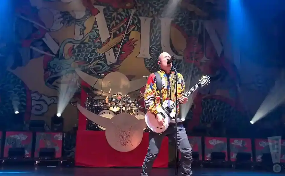 A bald man in a colorful jacket plays a white guitar on stage, with a drummer in the background.
