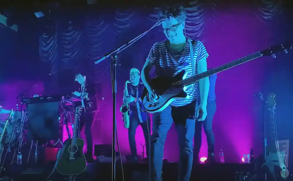 A band plays on a stage with purple and blue lighting. The lead singer plays a bass guitar, and a saxophonist is behind him.