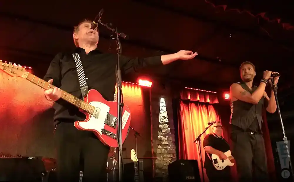 A male guitarist with a red guitar sings into a microphone on stage.