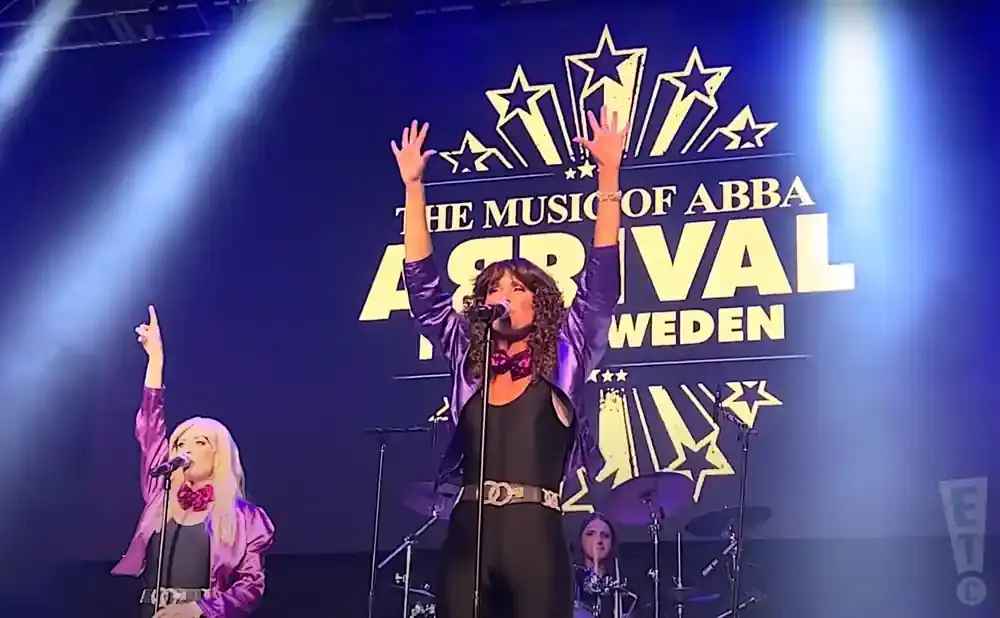 Two female singers perform on stage with a banner reading 