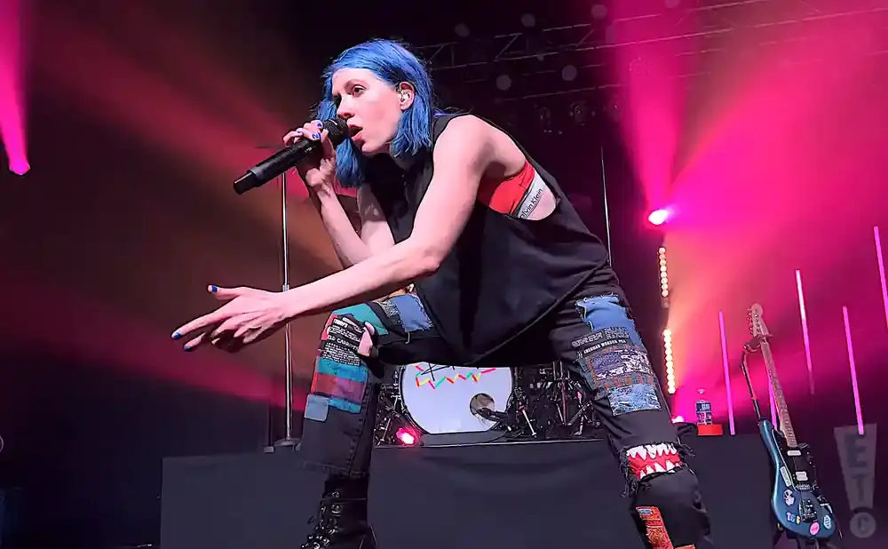 A woman with blue hair sings into a microphone on stage with pink lights.