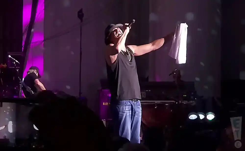 A male singer in a black tank top and jeans holds a white towel up in the air while singing into a microphone on stage.