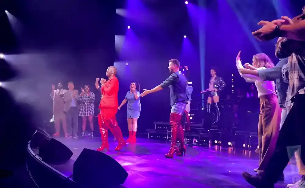 People on a stage in red boots and colorful outfits, some cheering.