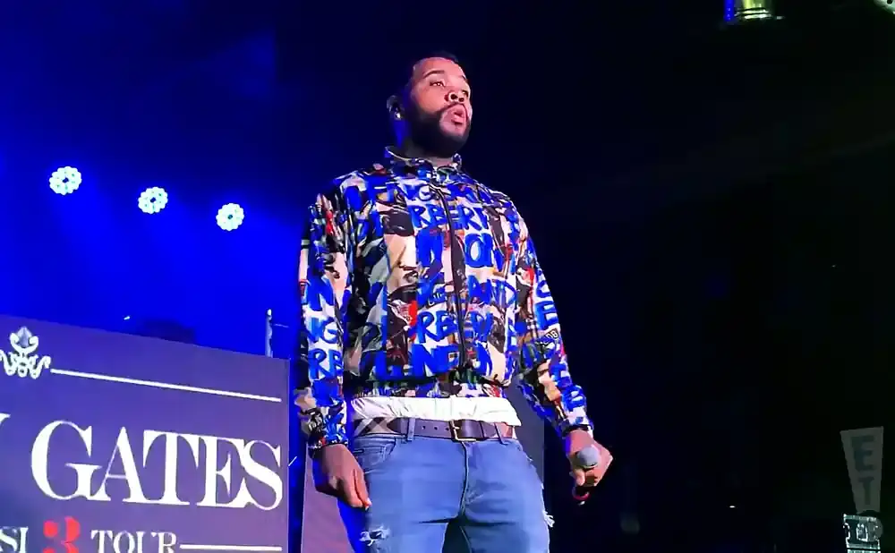 A man wearing a colorful jacket and jeans stands on stage under blue lights, holding a microphone.