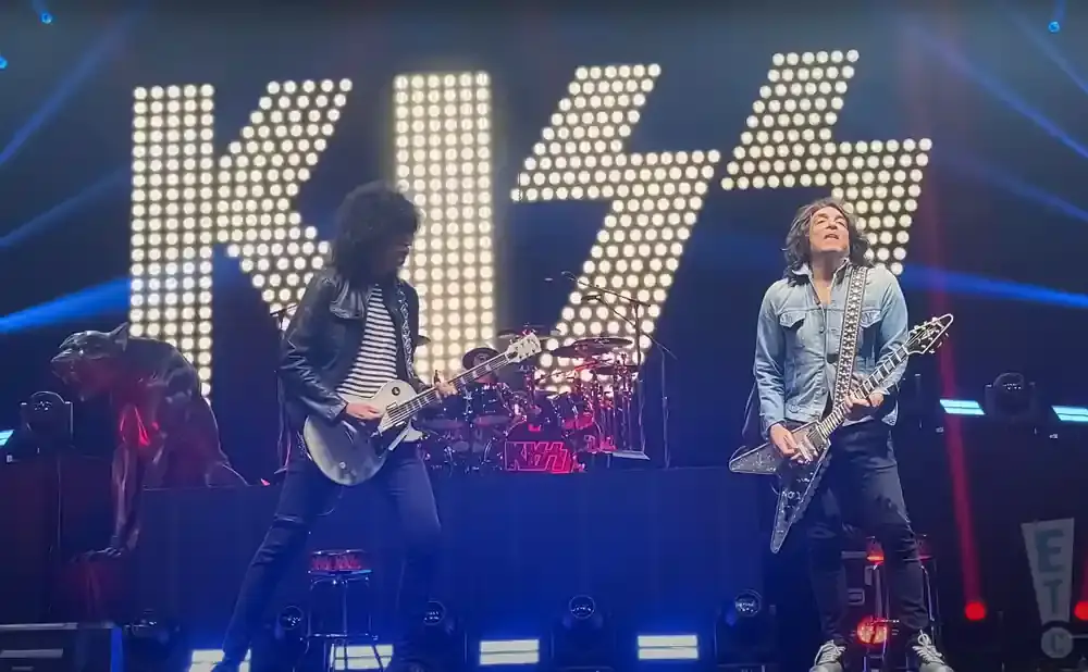 KISS band members Paul Stanley and Tommy Thayer play guitars on stage in front of a large, illuminated KISS logo.