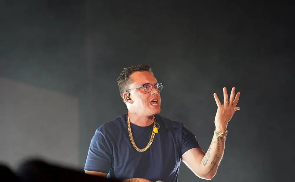 Rapper Logic performs on stage, wearing glasses, a gold chain, and a dark blue t-shirt, holding a microphone.