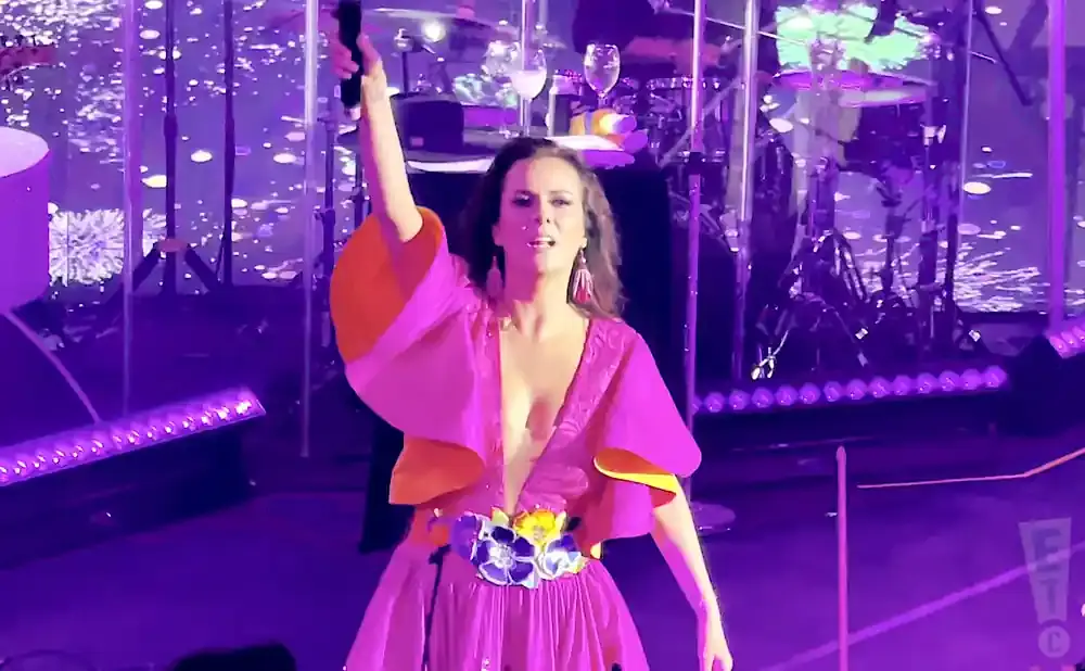 A woman in a pink and orange dress sings into a microphone on a stage with purple lights.