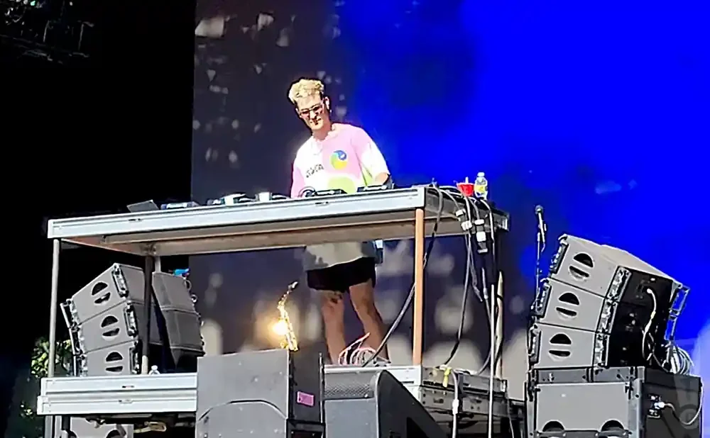 DJ performing on stage with speakers and equipment, wearing a colorful t-shirt.