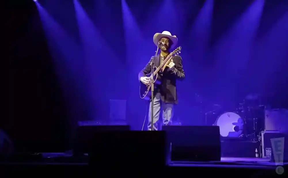 A man in a cowboy hat plays an acoustic guitar on a dimly lit stage, with blue spotlights overhead.
