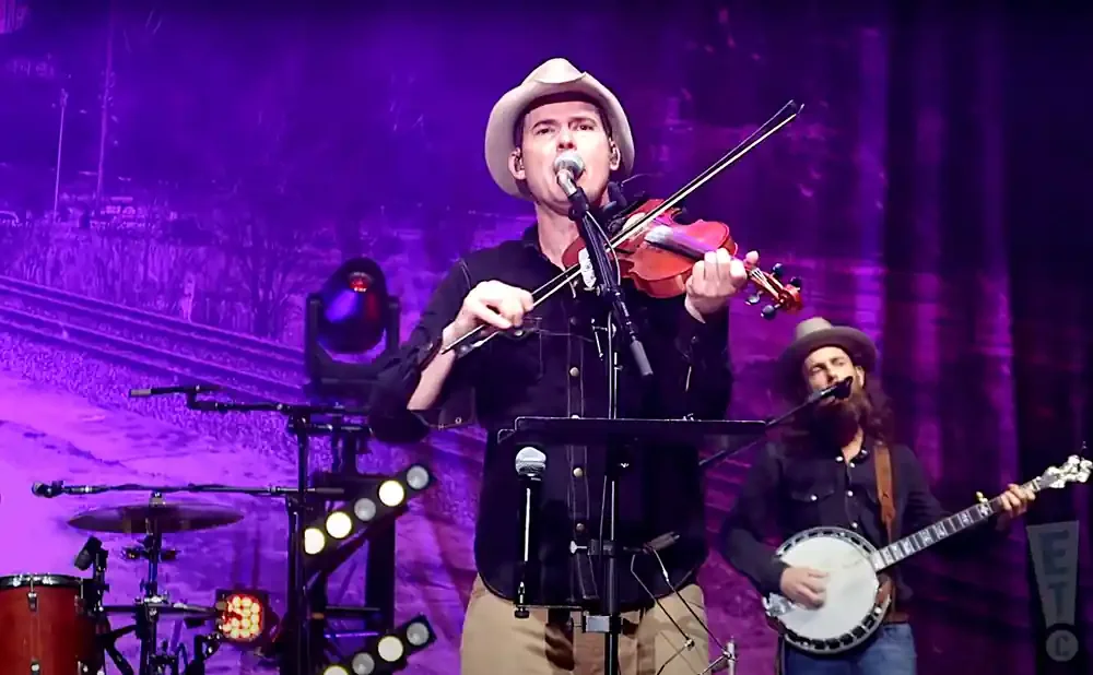 A man in a cowboy hat plays a violin on stage with a purple backdrop.