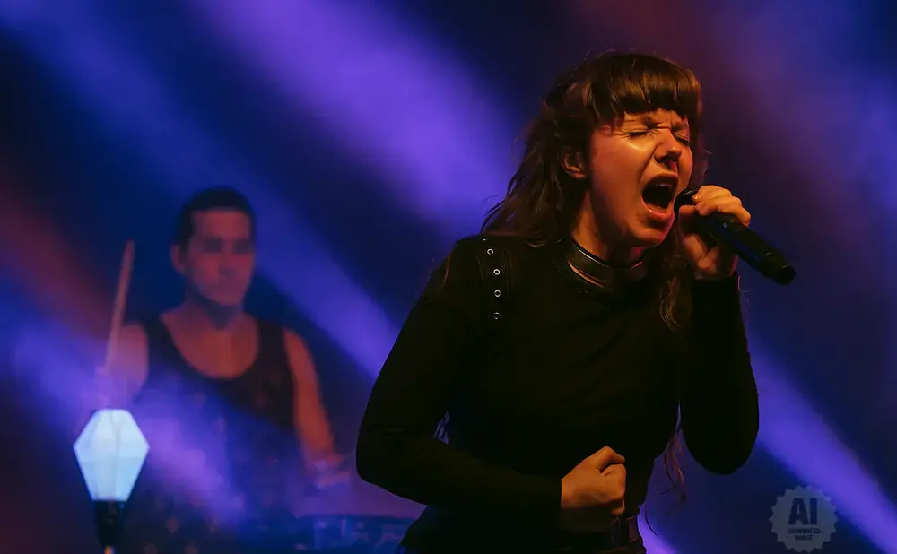A woman with her mouth open sings into a microphone on stage with a drummer in the background.