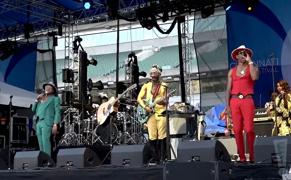 Musicians perform on a stage during a festival, with a banner in the background reading 