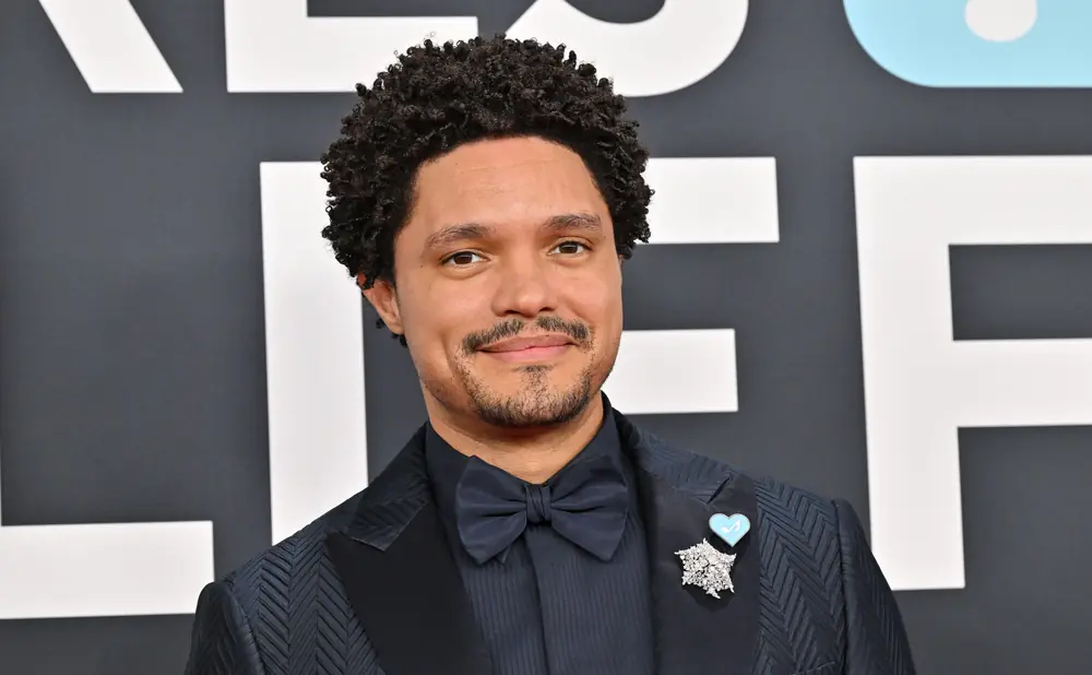 Trevor Noah smiles at the camera wearing a dark suit, bow tie, and a star brooch with a blue heart pin.