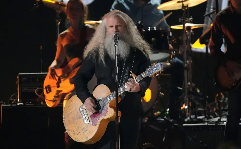 Musician with long white hair and beard plays a guitar covered in signatures while on stage.
