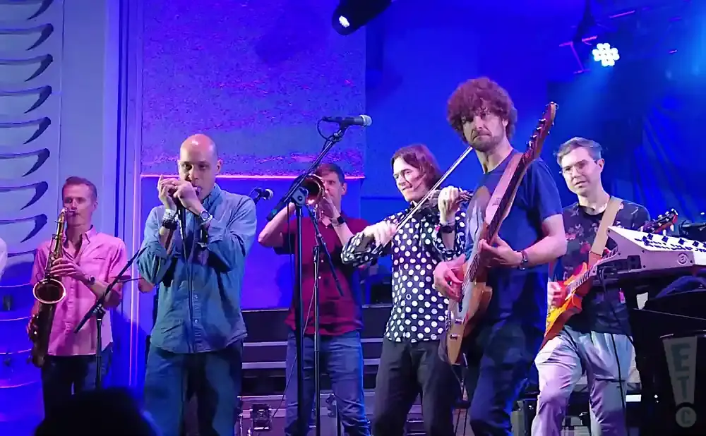 A band performs on stage with a saxophonist, harmonica player, trumpeter, violinist, guitarist, and keyboardist.