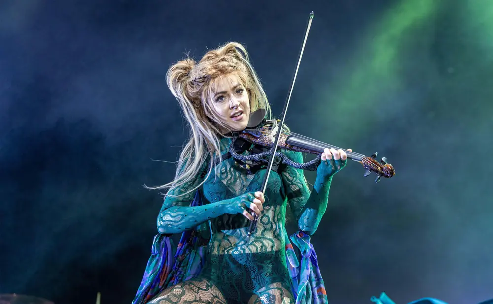 A woman in a green lace outfit plays an electric violin with a sparkly neck on a stage.