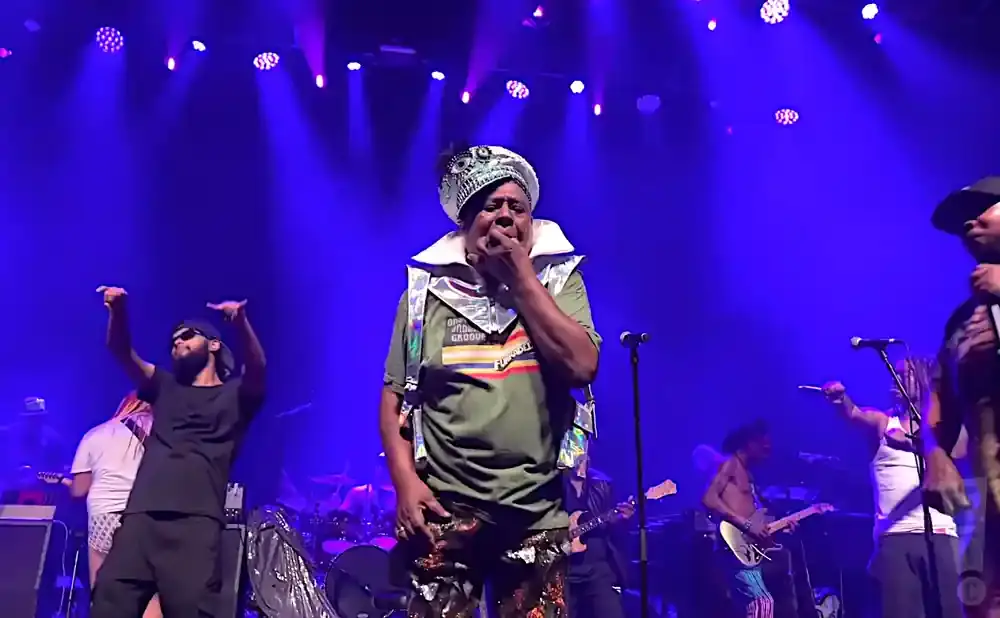 A band performs on a stage with blue lighting. The lead singer wears a sequined hat and outfit.