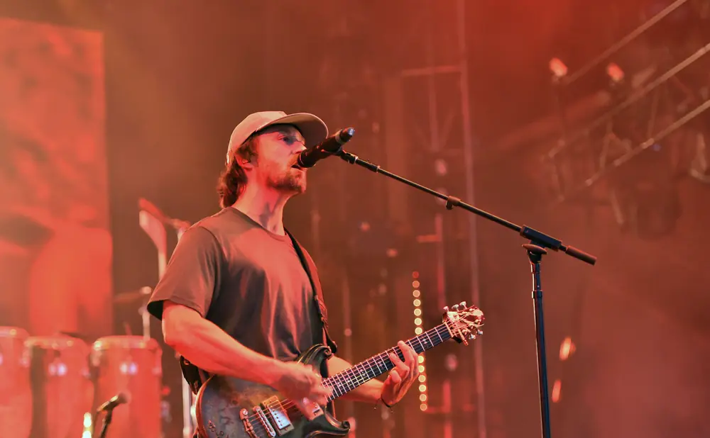 A male guitarist in a cap and t-shirt sings into a microphone while playing a dark electric guitar on stage.