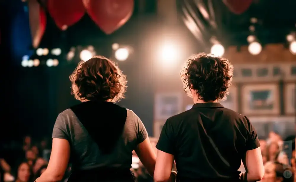 Two people with curly hair stand with their backs to the camera, facing a bright spotlight on a stage.