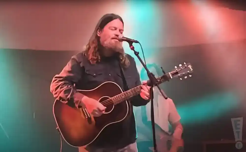 Musician with long hair and beard plays an acoustic guitar and sings into a microphone on a stage with colored lights.