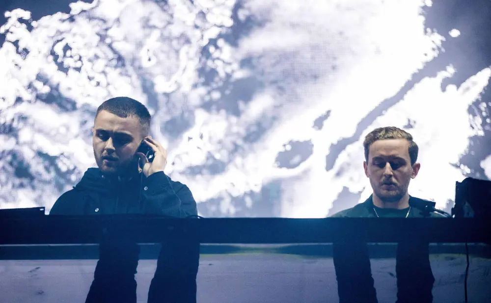 Two men stand behind a DJ booth with a large, abstract image projected behind them.