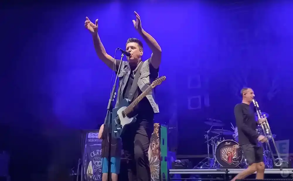 A man with a guitar sings into a microphone on a stage lit by blue lights, while another man plays a trombone.