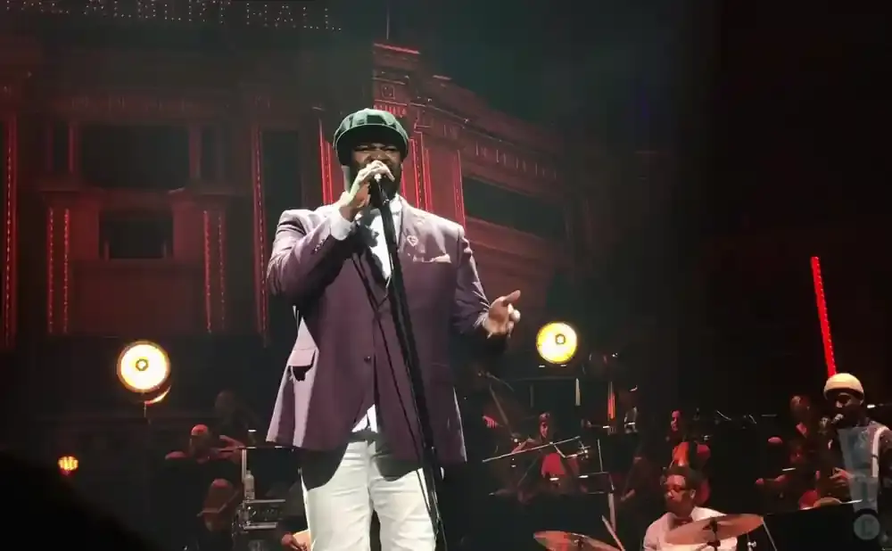 A singer in a purple suit and green cap performs on stage with an orchestra.