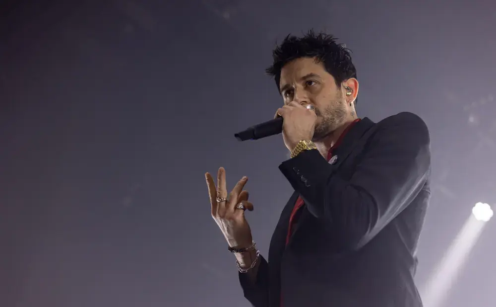 A man in a black suit and red shirt sings into a microphone, gesturing with his hands.
