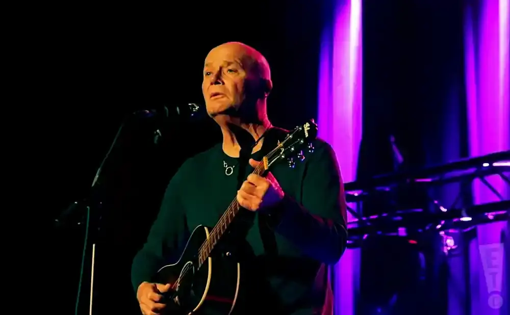 A bald man in a dark green sweater plays an acoustic guitar on stage with purple lights in the background.