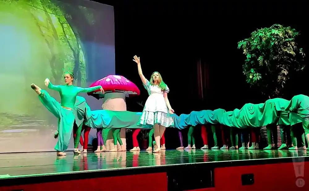 Dancers in a play perform on stage, with one dressed as a caterpillar and others as its segments, against a forest backdrop.