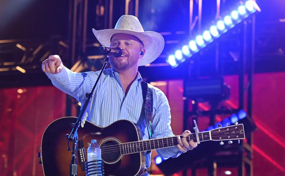 A man in a cowboy hat sings and plays an acoustic guitar on stage, illuminated by blue and red lights.