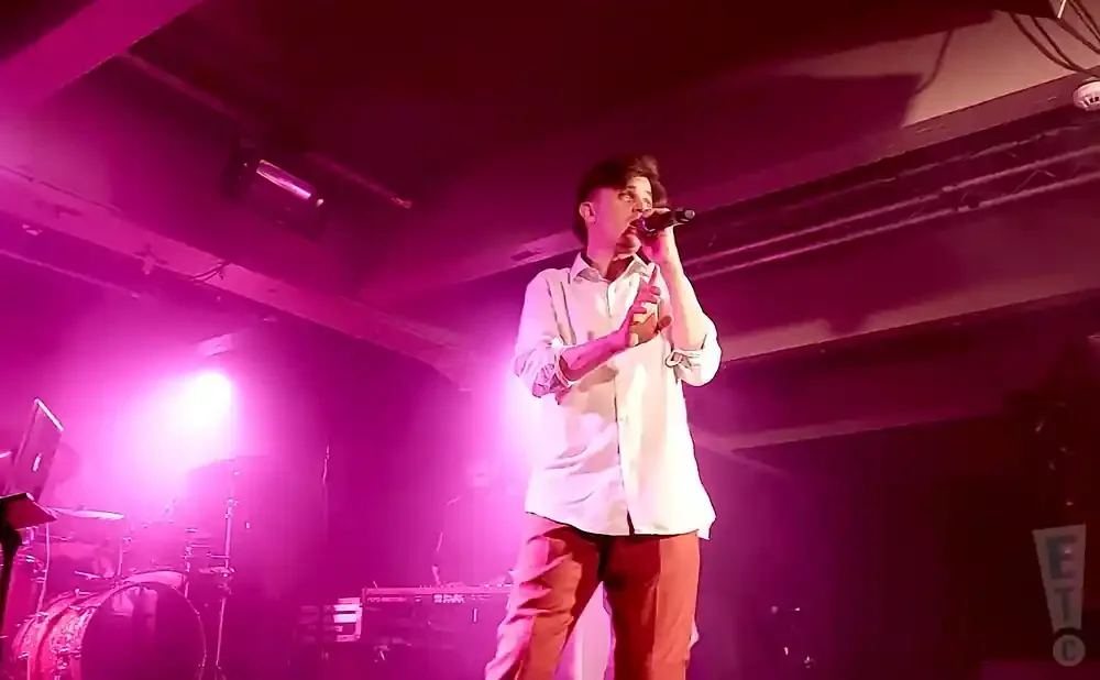 A male singer in a white shirt and orange pants performs on a dimly lit stage with pink lighting.