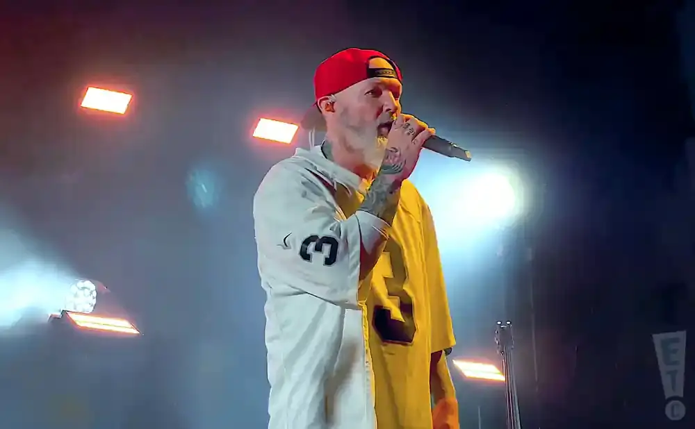 A tattooed man in a red baseball cap and a white and yellow shirt sings into a microphone on a stage with bright lights.