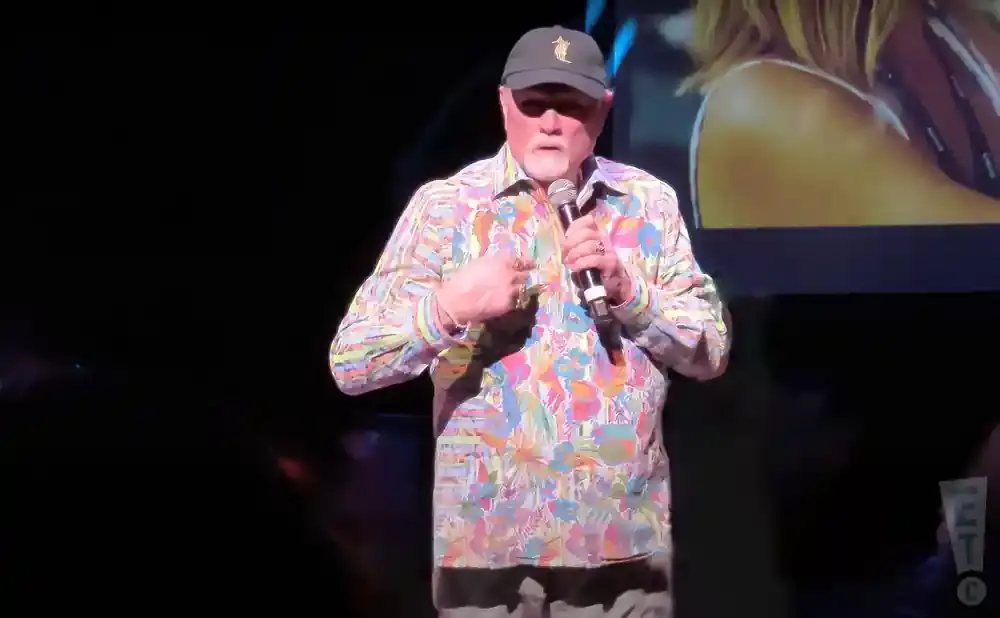 A man wearing a colorful, floral-patterned shirt and a baseball cap holds a microphone on stage.