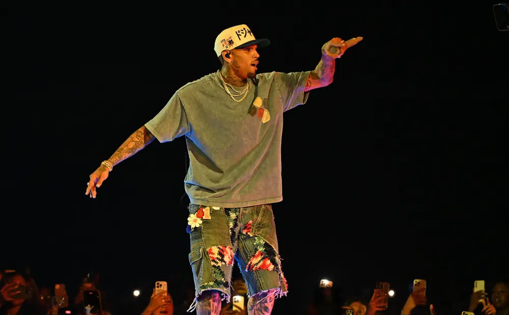 Chris Brown performs on stage, wearing a grey t-shirt, distressed denim shorts, and a baseball cap, with a crowd holding up phones in the foreground.