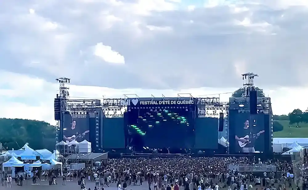 Large outdoor music festival stage with screens showing a performer playing guitar. A large crowd is gathered in front of the stage.