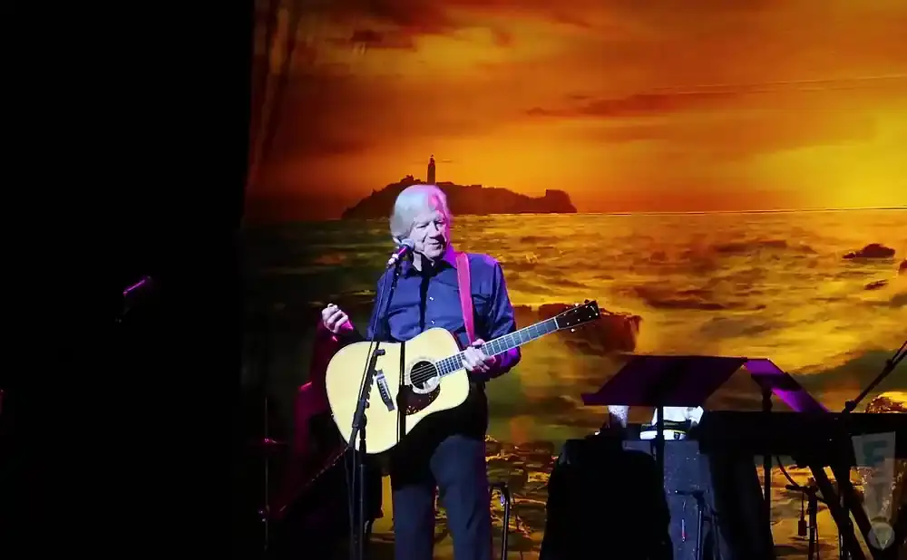 A man plays an acoustic guitar on stage in front of a sunset ocean backdrop.
