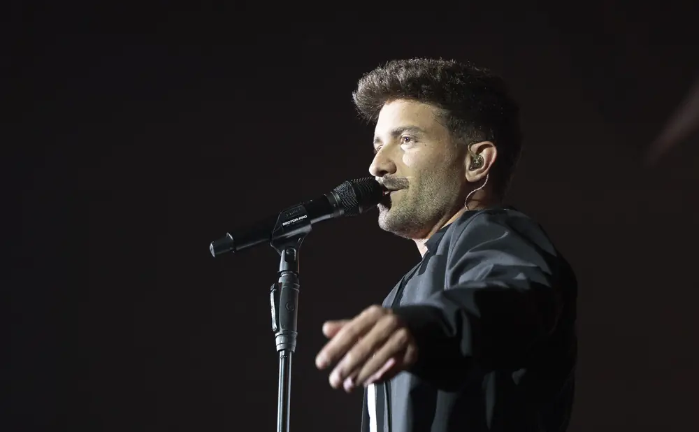 A man with dark hair sings into a microphone on stage.