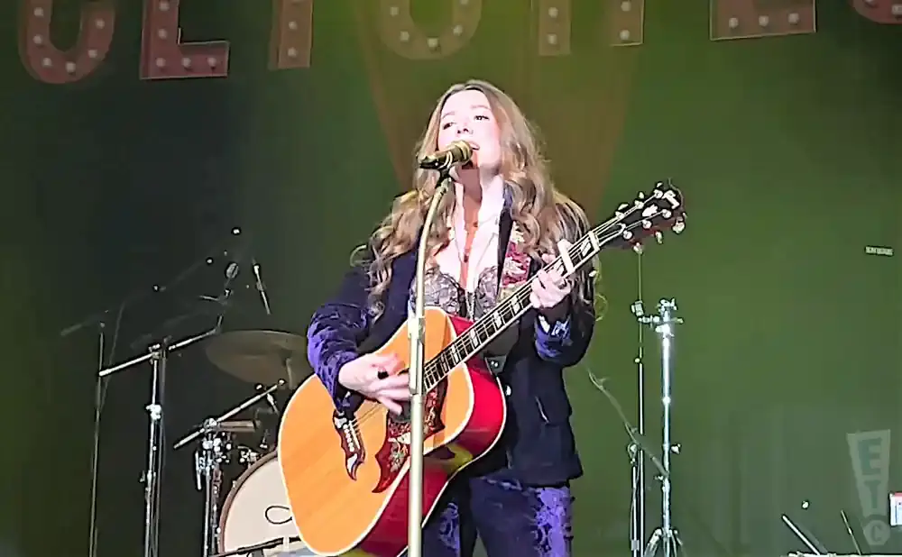 A woman in a purple suit plays an acoustic guitar and sings into a microphone on stage.