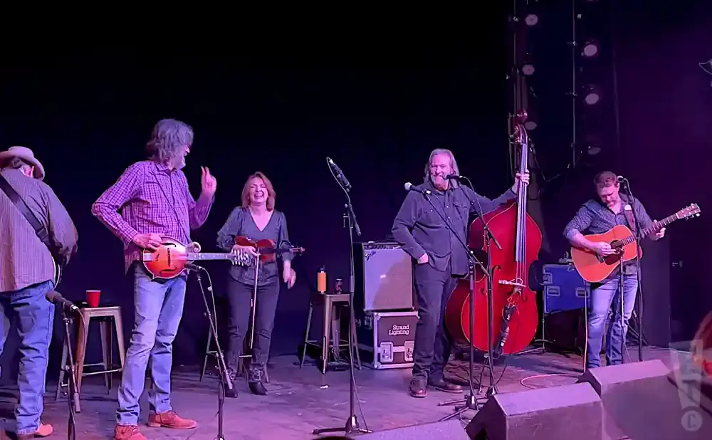 A band plays music on a stage with a dark background. From left to right, a man in a hat plays guitar, a man plays mandolin, a woman plays violin, a man…