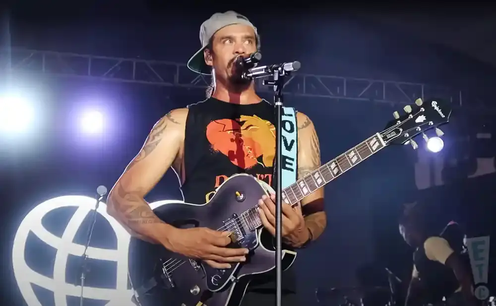Musician with tattoos, wearing a baseball cap and a tank top with a heart design, plays an electric guitar on stage.