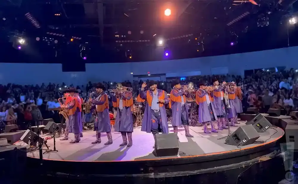 A band in matching purple and orange outfits performs on stage with trumpets and saxophones in front of a large audience.