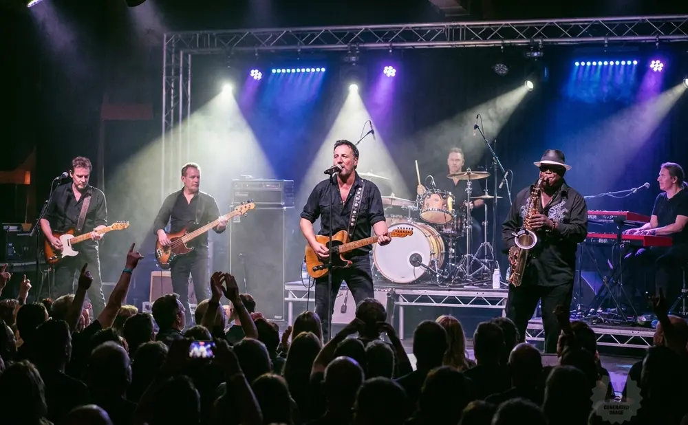 A band performs on stage with a saxophonist, guitarist, drummer, and keyboardist, lit by purple and white spotlights.
