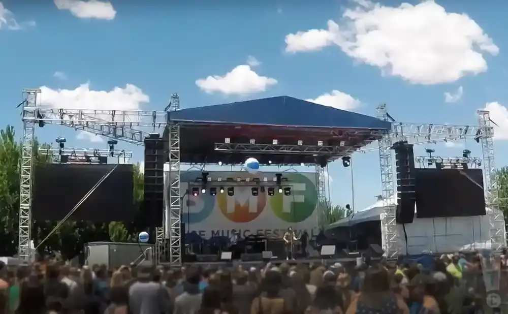 A live music festival stage with screens, speakers, and a crowd in front.