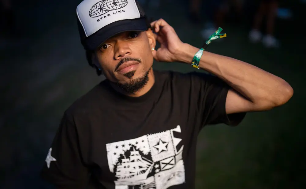 A man with a goatee wears a black t-shirt with a white graphic and a trucker hat.
