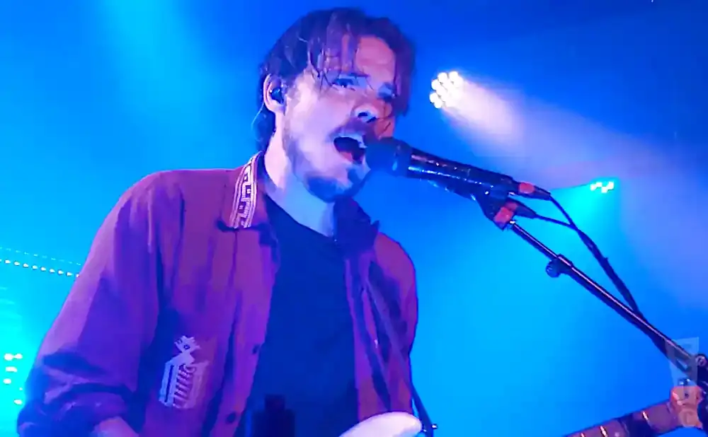 A man with dark hair sings into a microphone on a blue-lit stage, wearing a dark shirt and a maroon jacket.
