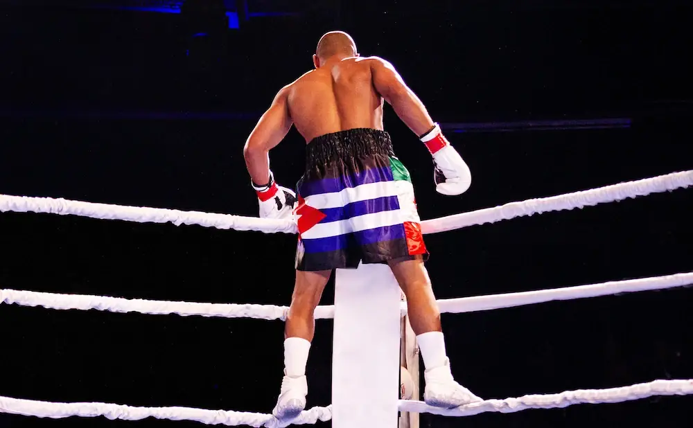 Boxer in colorful shorts and white gloves stands in a boxing ring.