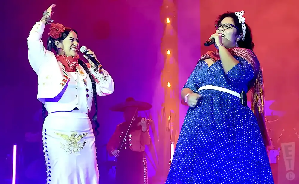 Two women in colorful outfits sing on stage with a violinist in the background.